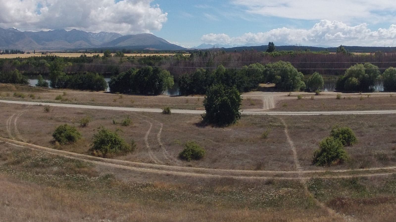 Open ground and gravel access tracks at Lake Poaka Campsite, with trees along the water and distant mountains.