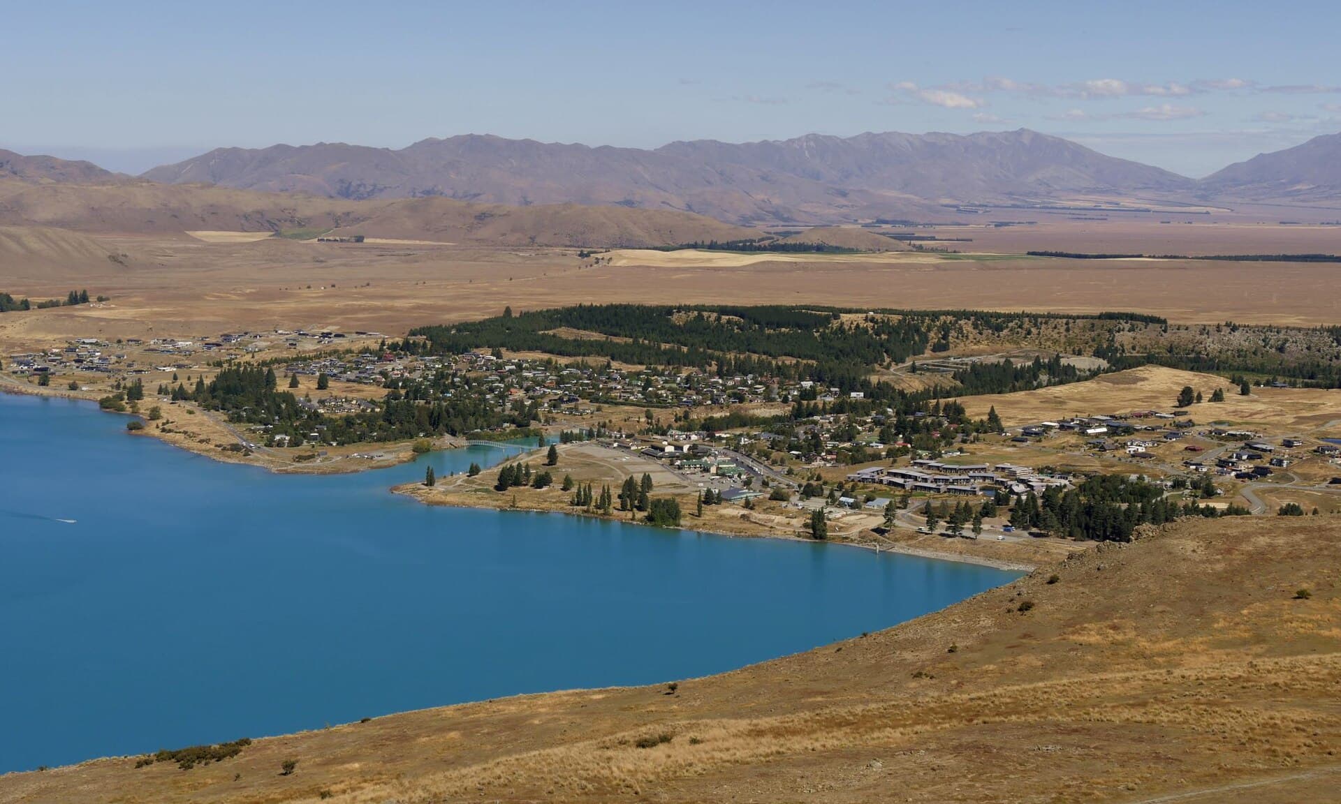 Lake Tekapo township along the blue lake, with dry open land and mountains behind, viewed from Mount John.