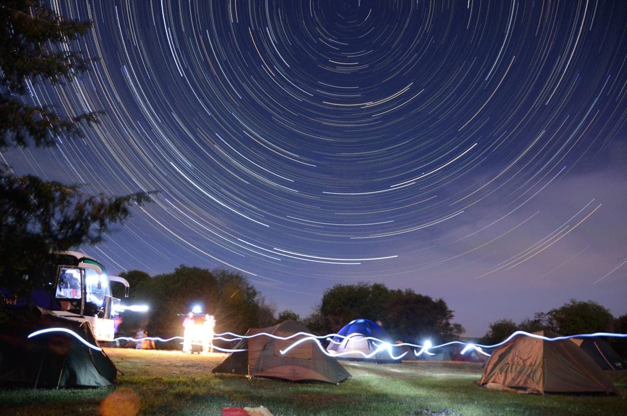 Several tents and a camping vehicle on grass at night beneath curved star trails at Lake Rerewhakaaitu.