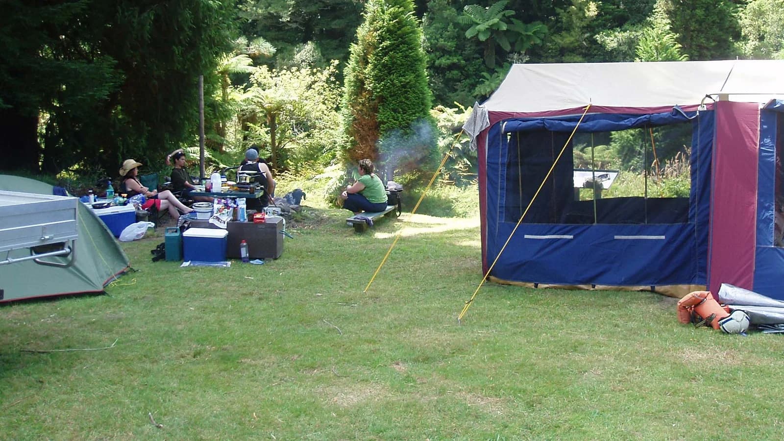 Tents, camping equipment and a seated group on a grassy pitch beside forest at Whitikau Campsite.