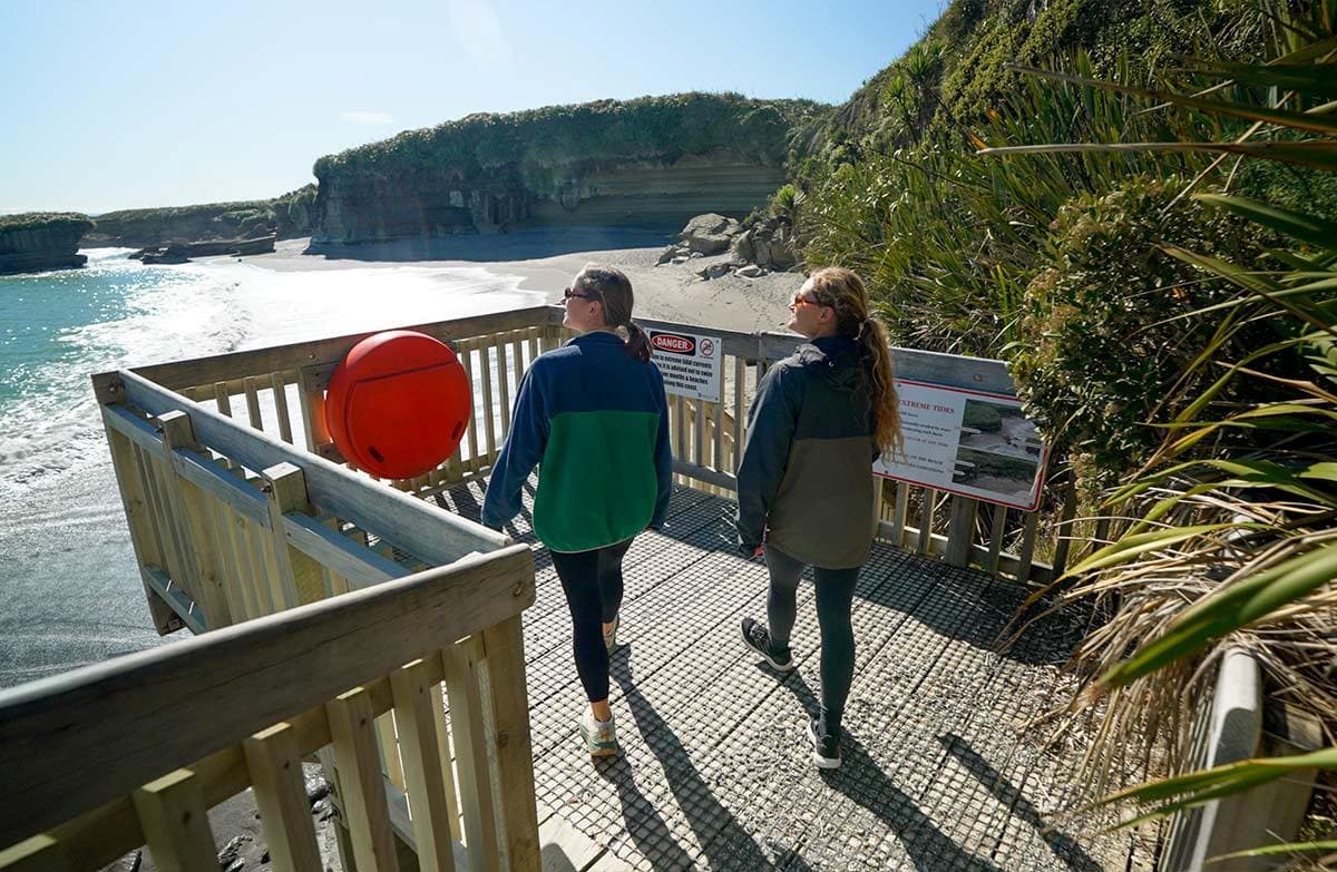 Two walkers on a wooden platform above a beach with sandstone cliffs, lush coastal vegetation and shining sea.