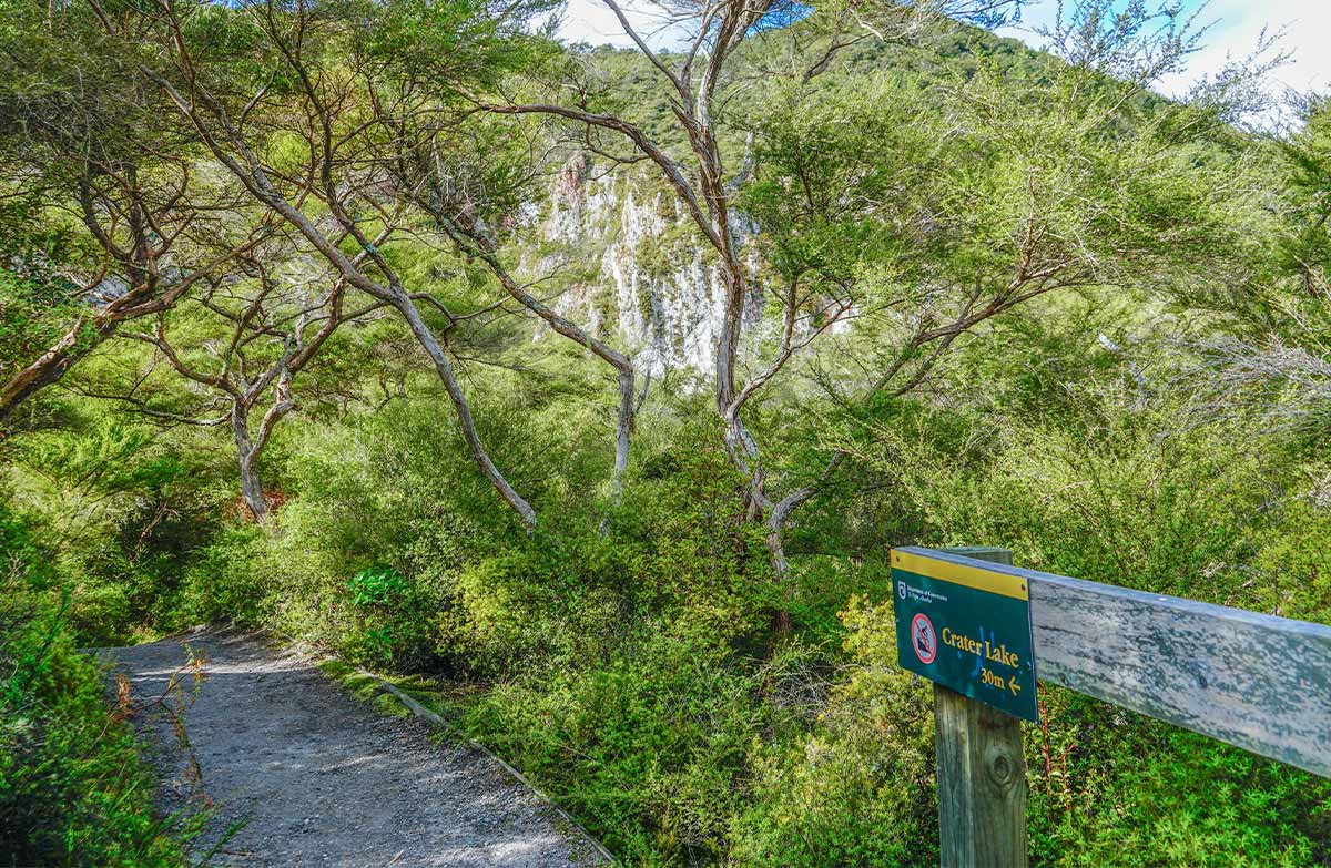 A gravel walking track through native bush, with a sign pointing to Crater Lake 30 metres ahead and rocky cliffs visible through the trees.