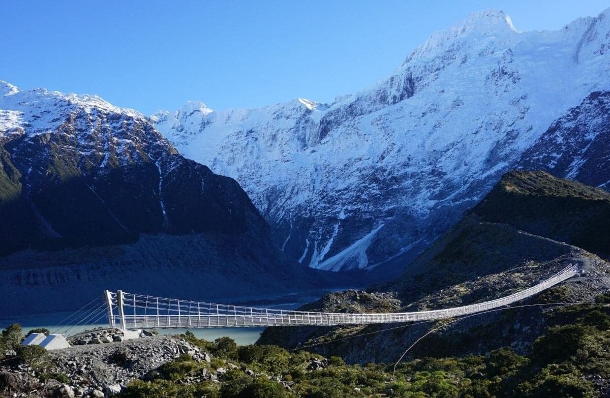  Suspension bridge beneath snow-covered mountains, with a glacial lake in the background.