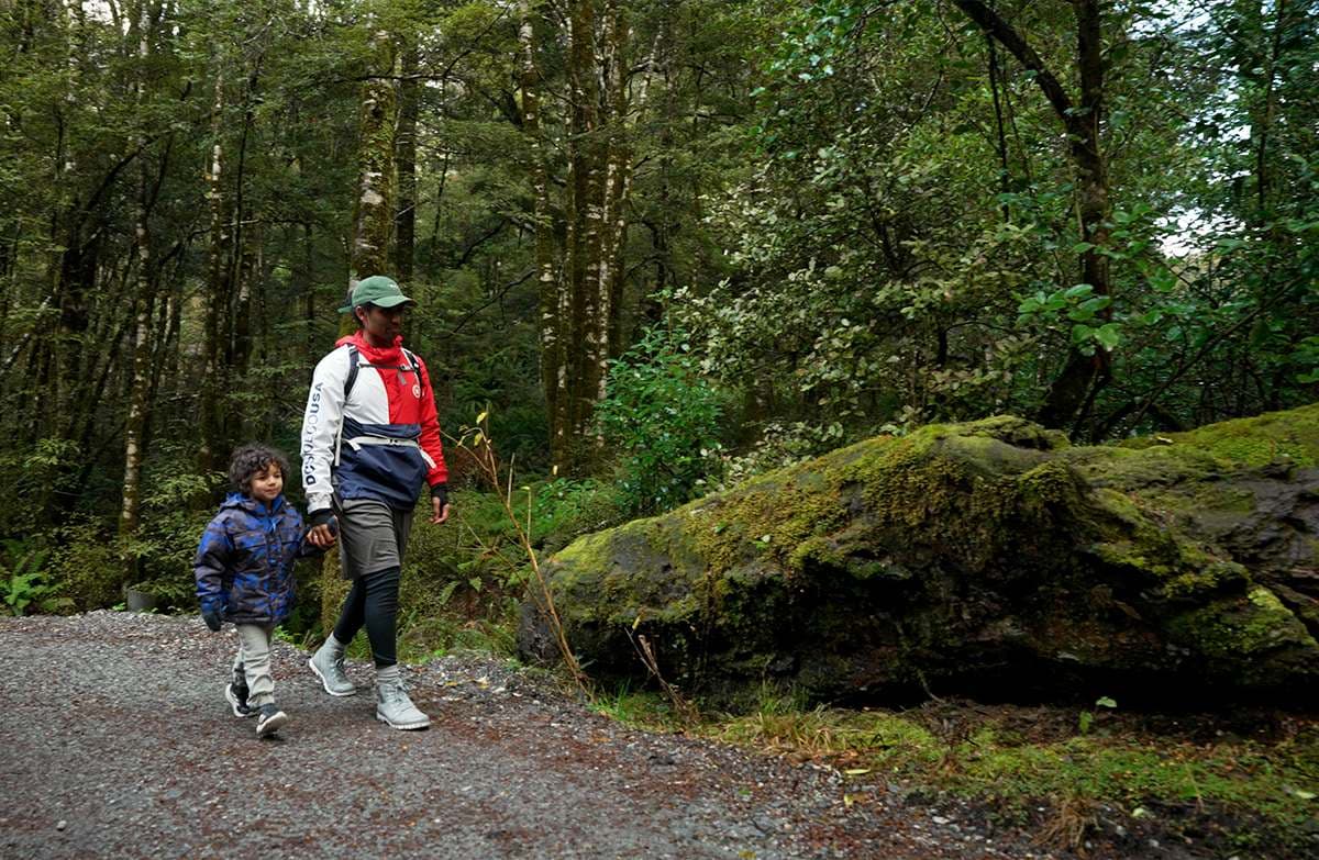An adult and child walk hand in hand along a gravel path past a large mossy rock.