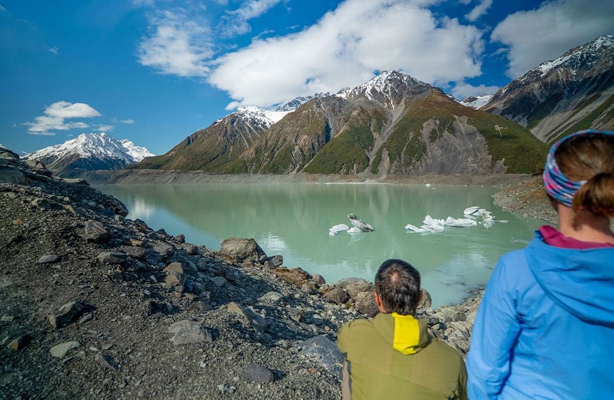 Two walkers face a milky turquoise lake with icebergs close to shore and snowy peaks beyond.