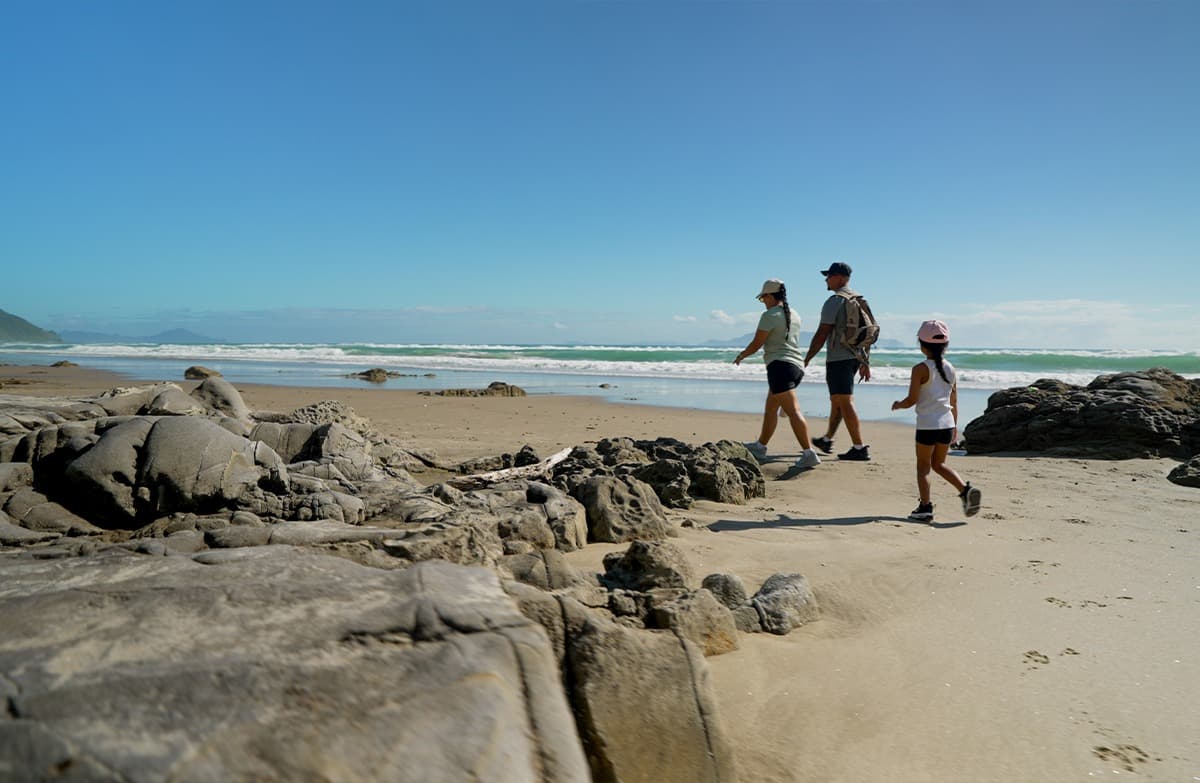 A family walking along a sandy beach with rocks and breaking waves.