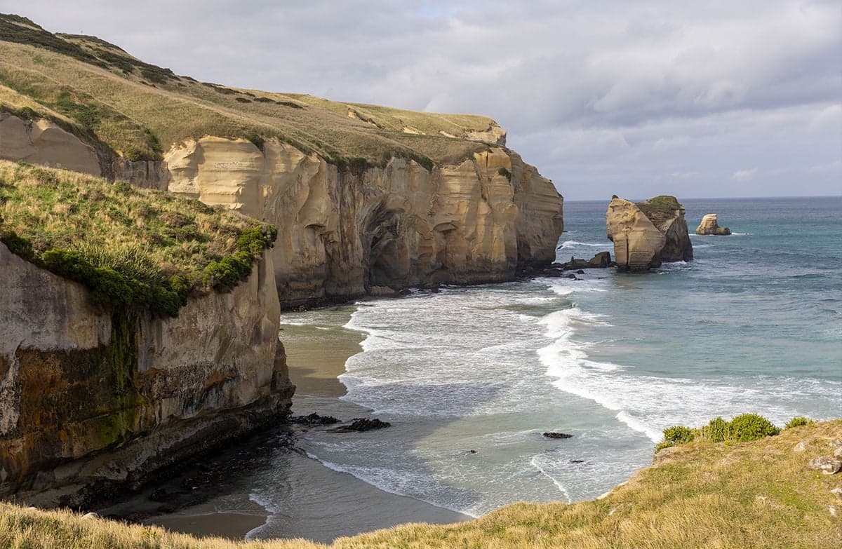 High sandstone cliffs covered in tussock tower above a secluded bay where waves are rolling ashore.