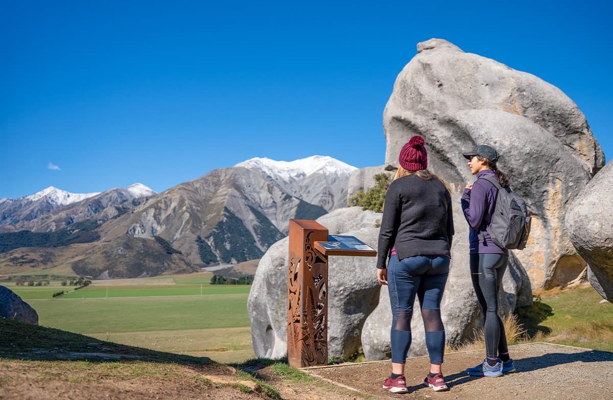 Two walkers beside boulders and a sign with Māori motifs look out to farmland and snowy mountains.