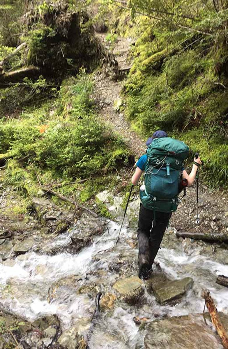 A person crosses a small stream by stepping on flat small rocks jutting out of the stream. The track is rough and steep.