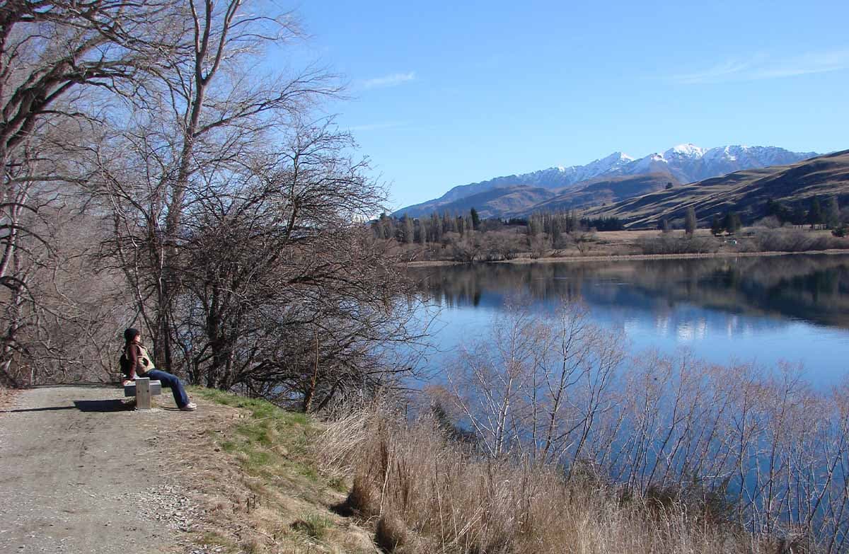 A person sits on a small wooden bench looking out across a serene lake. Mountains topped with snow can be seen far in the distance.