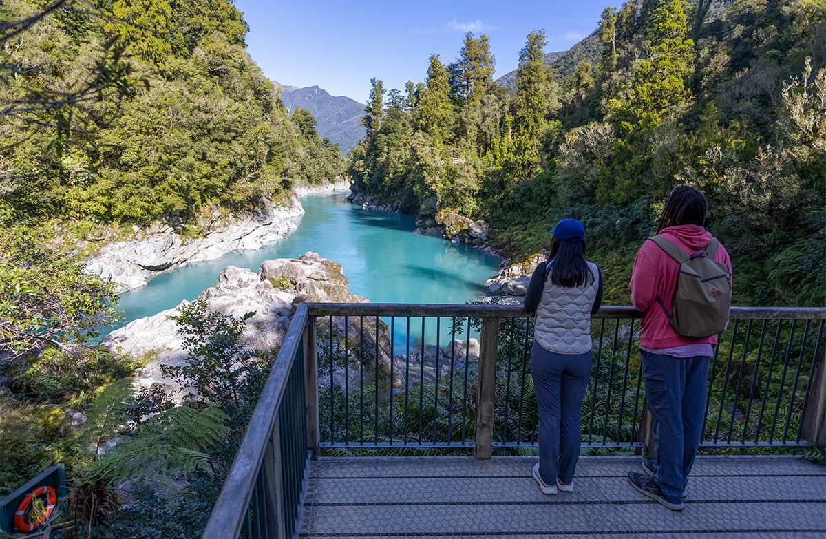 Two walkers on a platform admire a wide river gorge with turquoise water and tall trees.