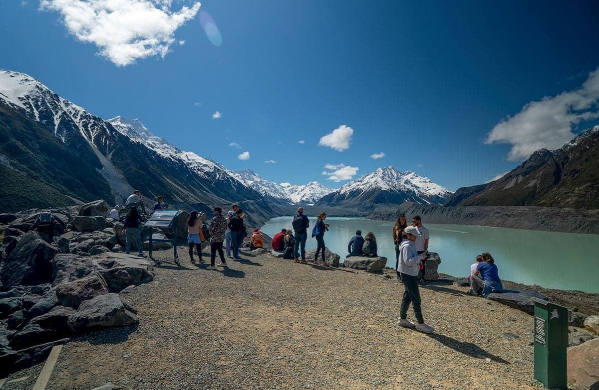 Walkers gather at a lookout above a glacial lake surrounded by snow-capped mountains.