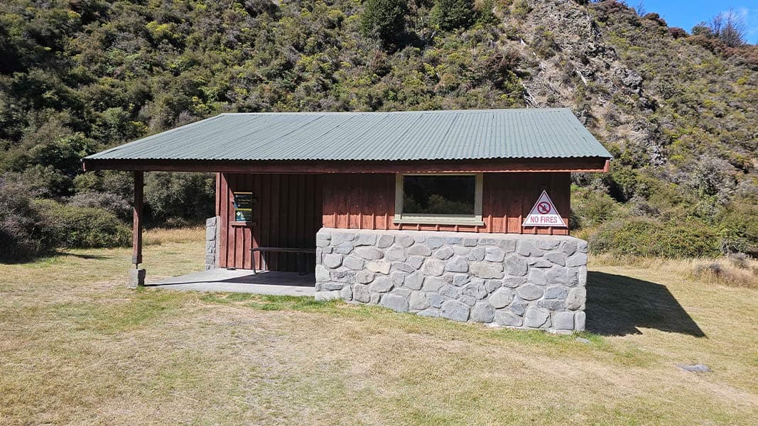 Timber shelter with a green roof, stone-faced wall and no-fires sign on grass at Andrews Shelter Campsite.