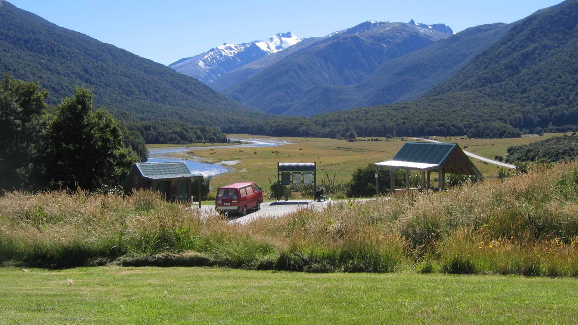 Grassy ground, a shelter, information board and red van at Cameron Flat, with a river and mountains behind.