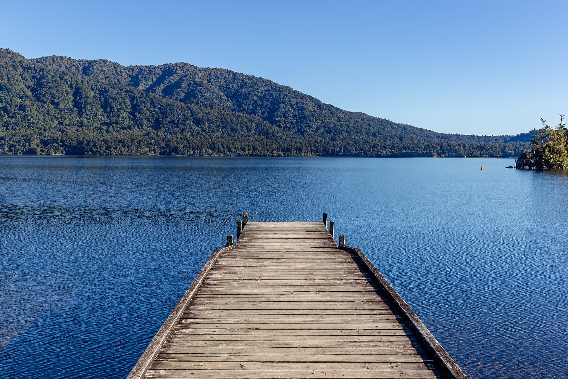 A timber pier extending into blue Lake Kaniere with forested hills along the opposite shore.