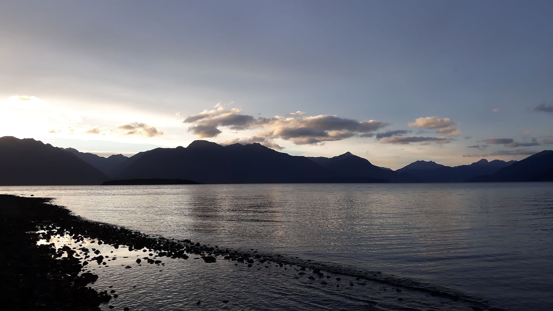 A stony shoreline and rippling Lake Te Anau beneath mountain silhouettes and an evening sky at Henry Creek.