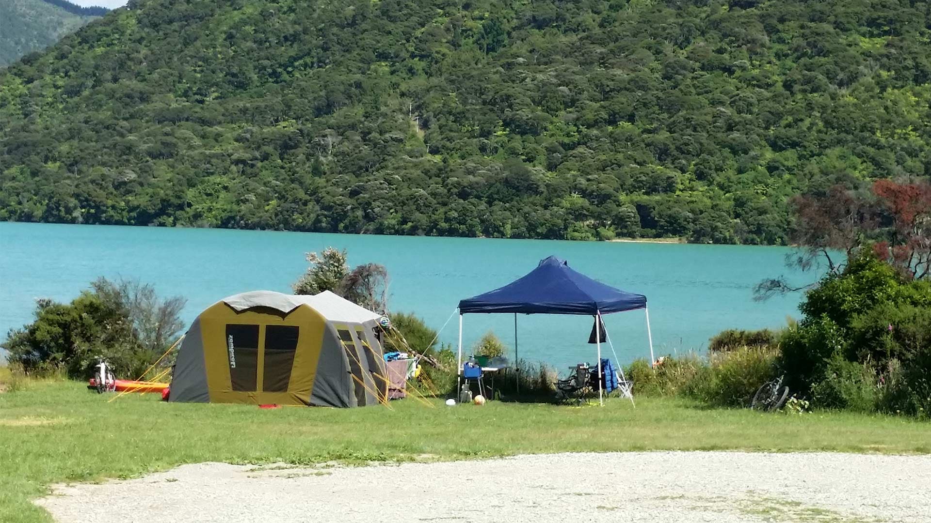Large tent and blue gazebo pitched on grass beside the water at Kenepuru Head Campsite.