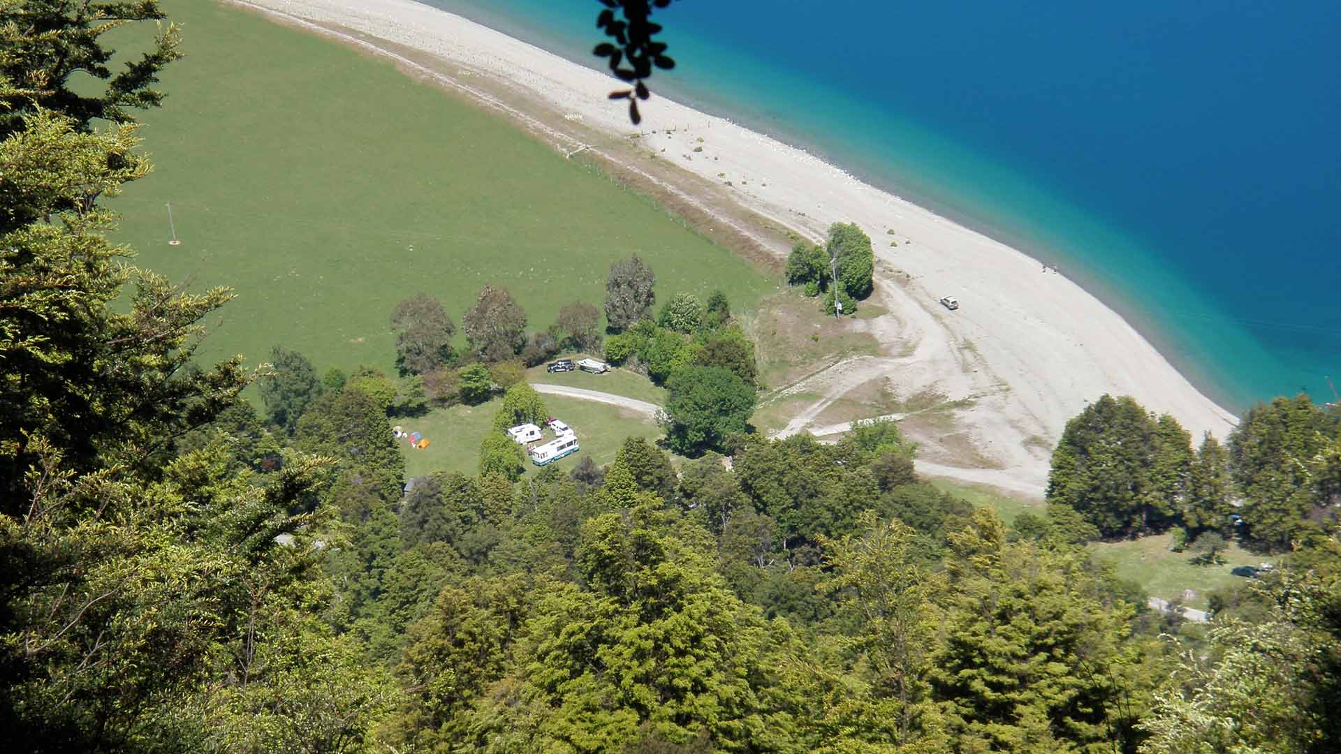 Tents and campervans among trees at Kidds Bush, seen from above beside the blue water of Lake Hawea.