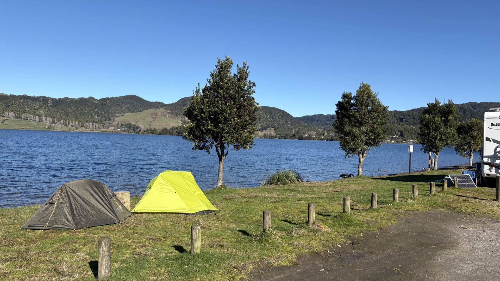 Two tents and a campervan on grassy ground beside Lake Okareka, with trees along the shore.
