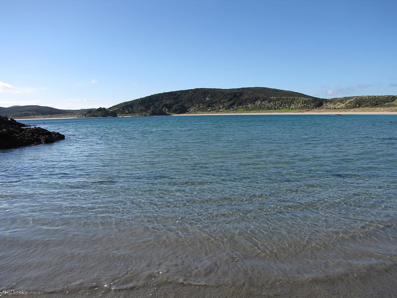Clear shallow water and a long sandy beach beneath low hills at Maitai Bay.
