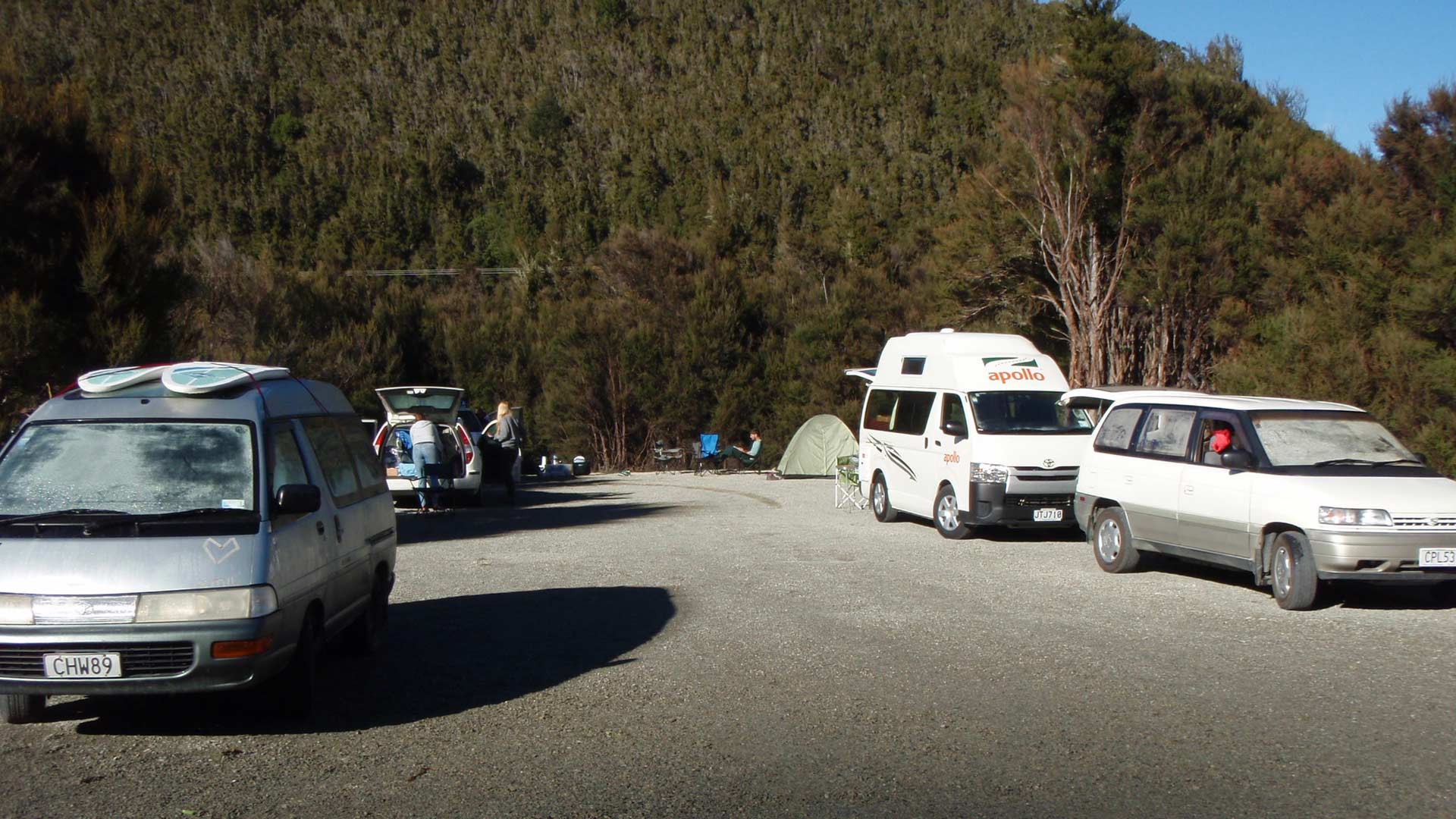 Campervans, cars and a small tent on gravel at Teetotal Campsite beside bush-covered slopes.