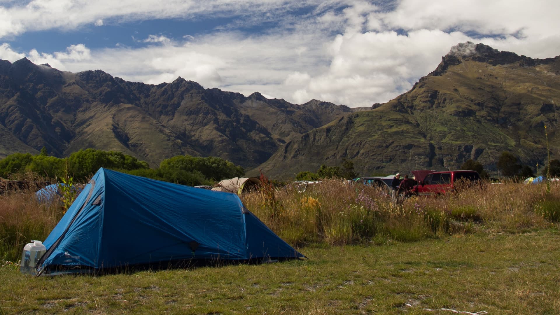 A blue tent in front of other tents and parked vehicles at Twelve Mile Delta, beneath rugged mountains.