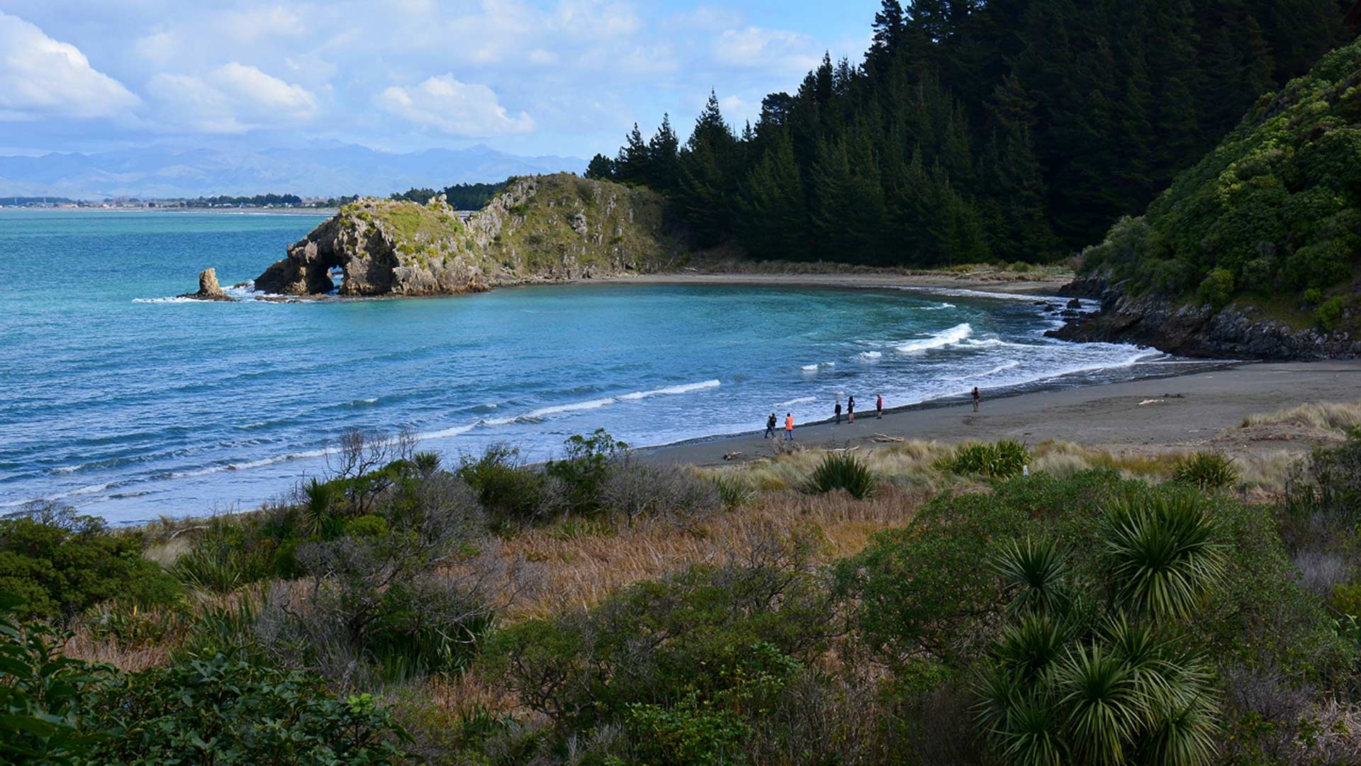 Curving sandy beach at Whites Bay with people on the shore, blue water and wooded hills.