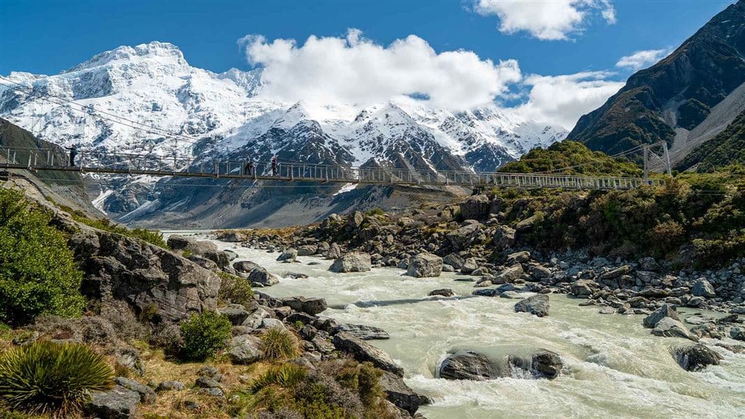A suspension bridge over the rocky Hooker River, backed by snow-covered mountains.