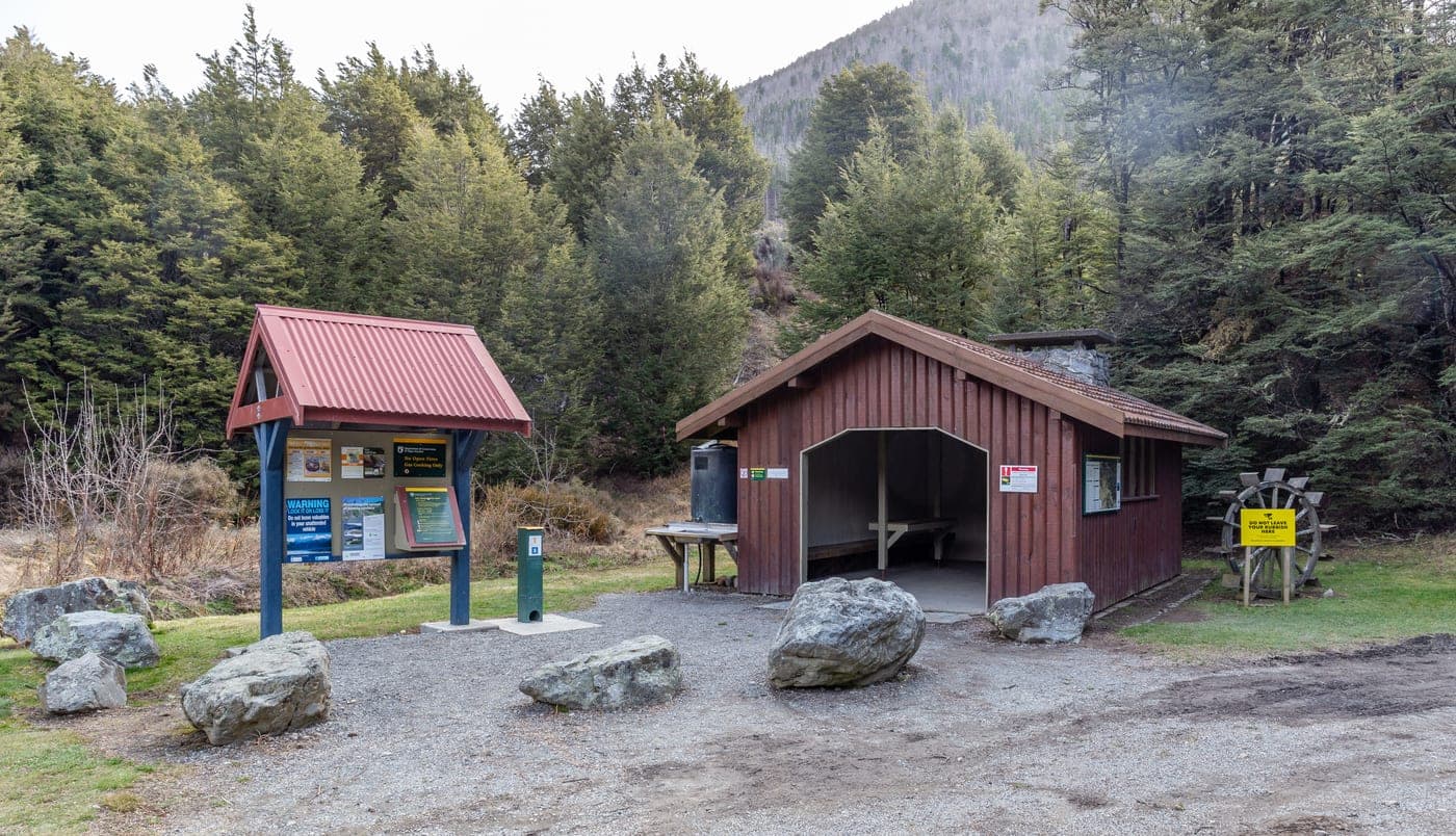 A small timber campsite shelter with a covered table, an outdoor sink and an information board beside forest.