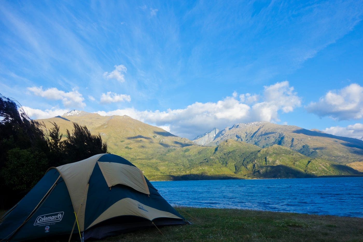 A blue and beige dome tent on grass beside Lake Hawea, with the lake and mountains beyond.