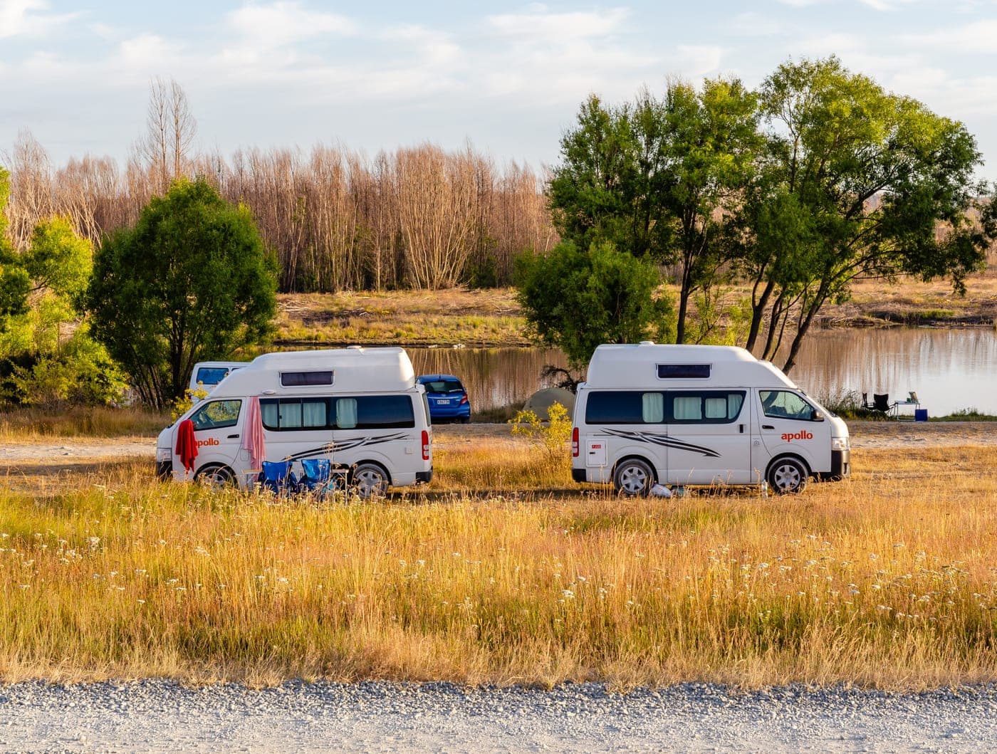Two white campervans and folding chairs on grassy ground beside trees and water at Lake Poaka.