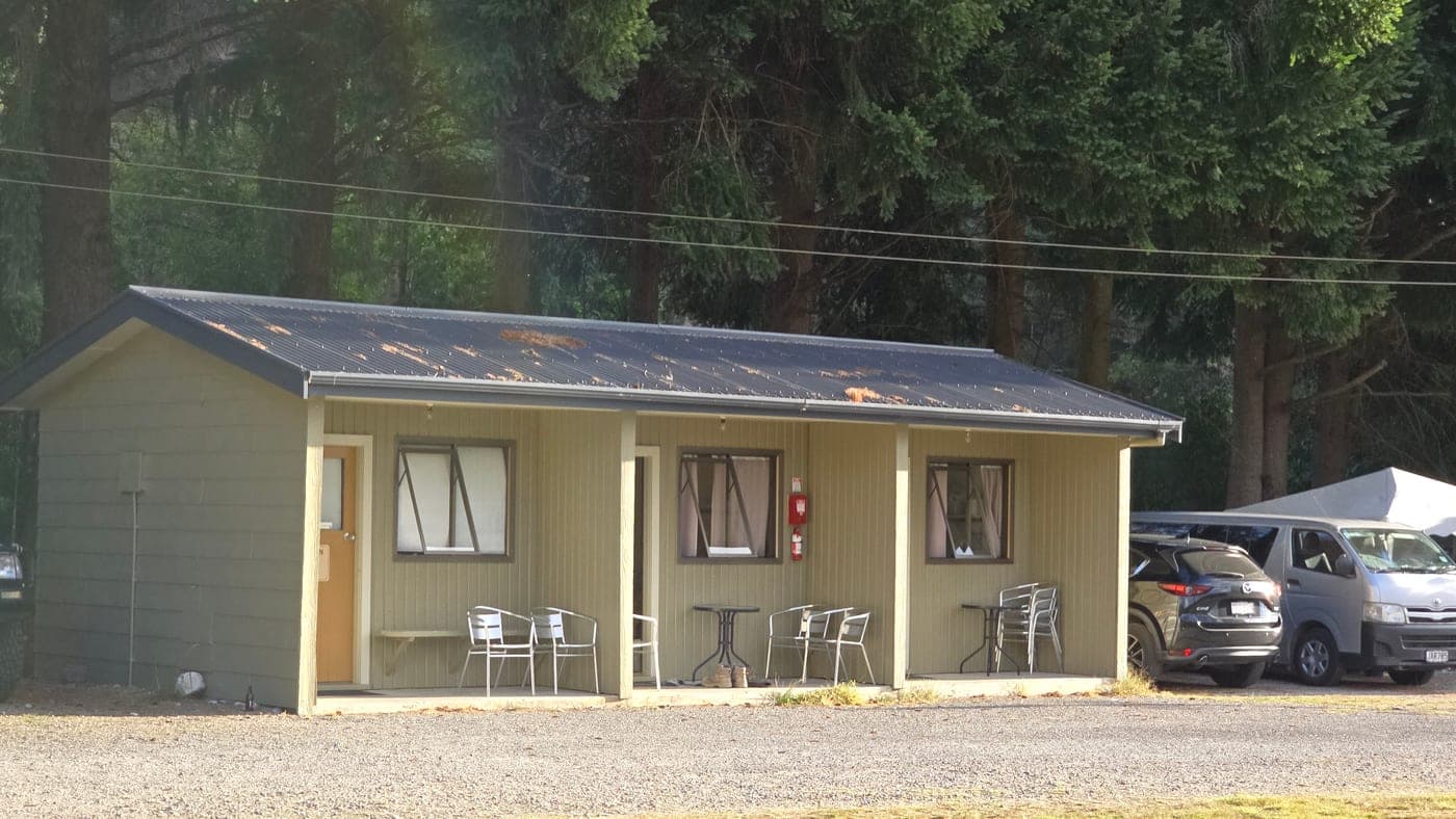 Small campground cabins with covered porches, outdoor chairs and tables, with vehicles and tall trees nearby.