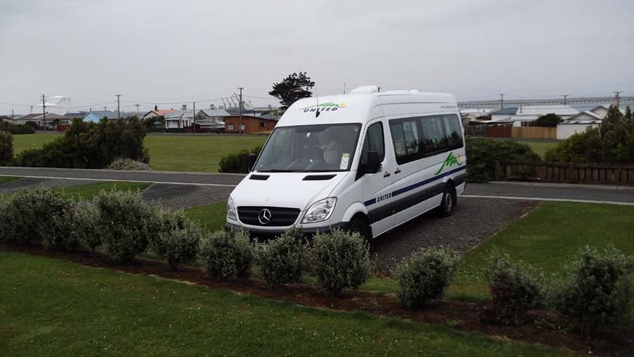 A campervan on a gravel pitch bordered by grass and shrubs at Bluff Camping Ground, with houses and port cranes behind; not Tasman Picton.