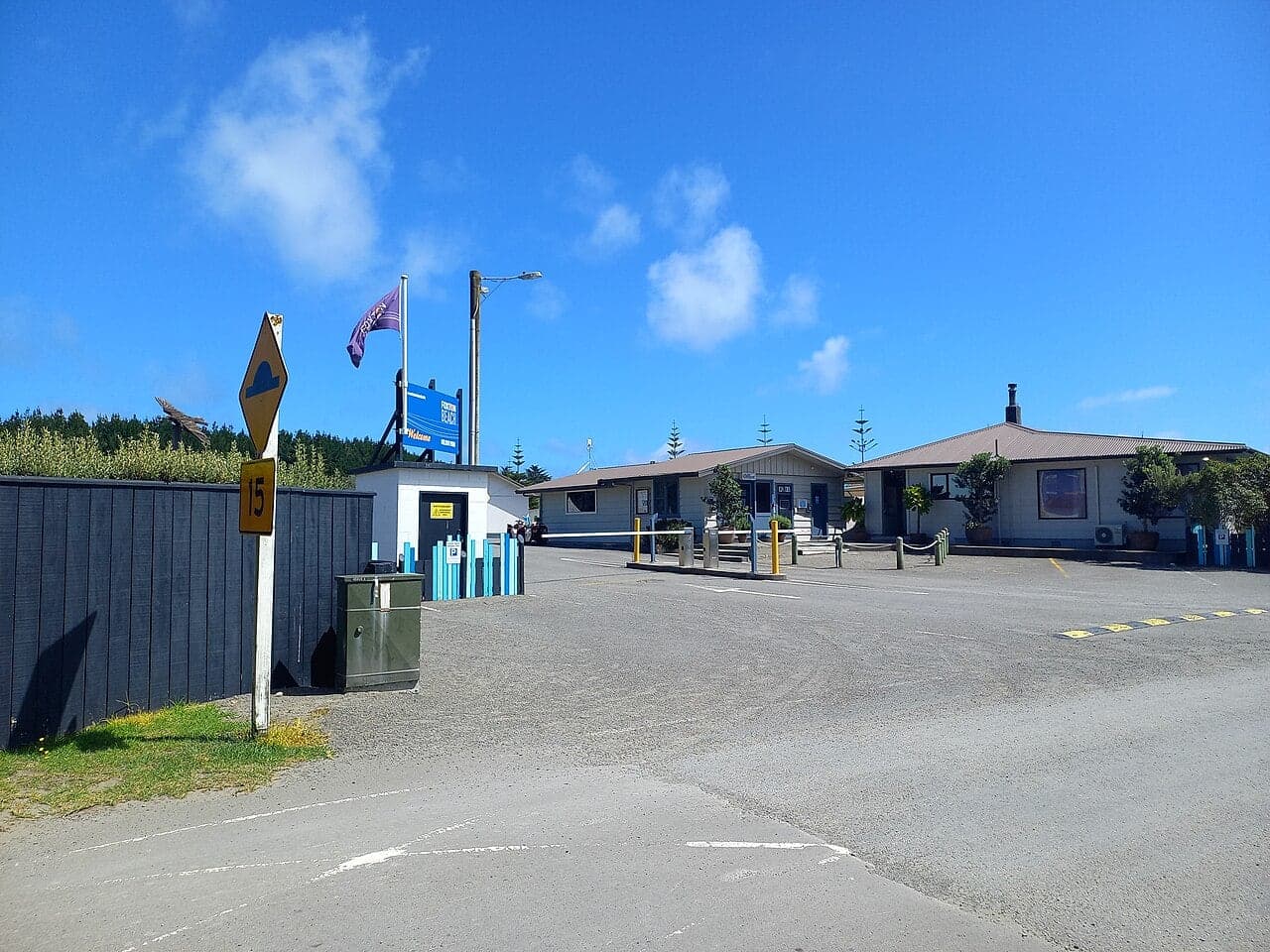 Foxton Beach Holiday Park entrance with a barrier arm, blue sign, reception buildings and paved driveway.
