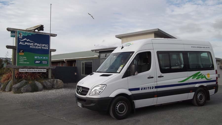 A campervan beside the Alpine-Pacific Holiday Park entrance sign, street number 69 and campground buildings in Kaikoura.