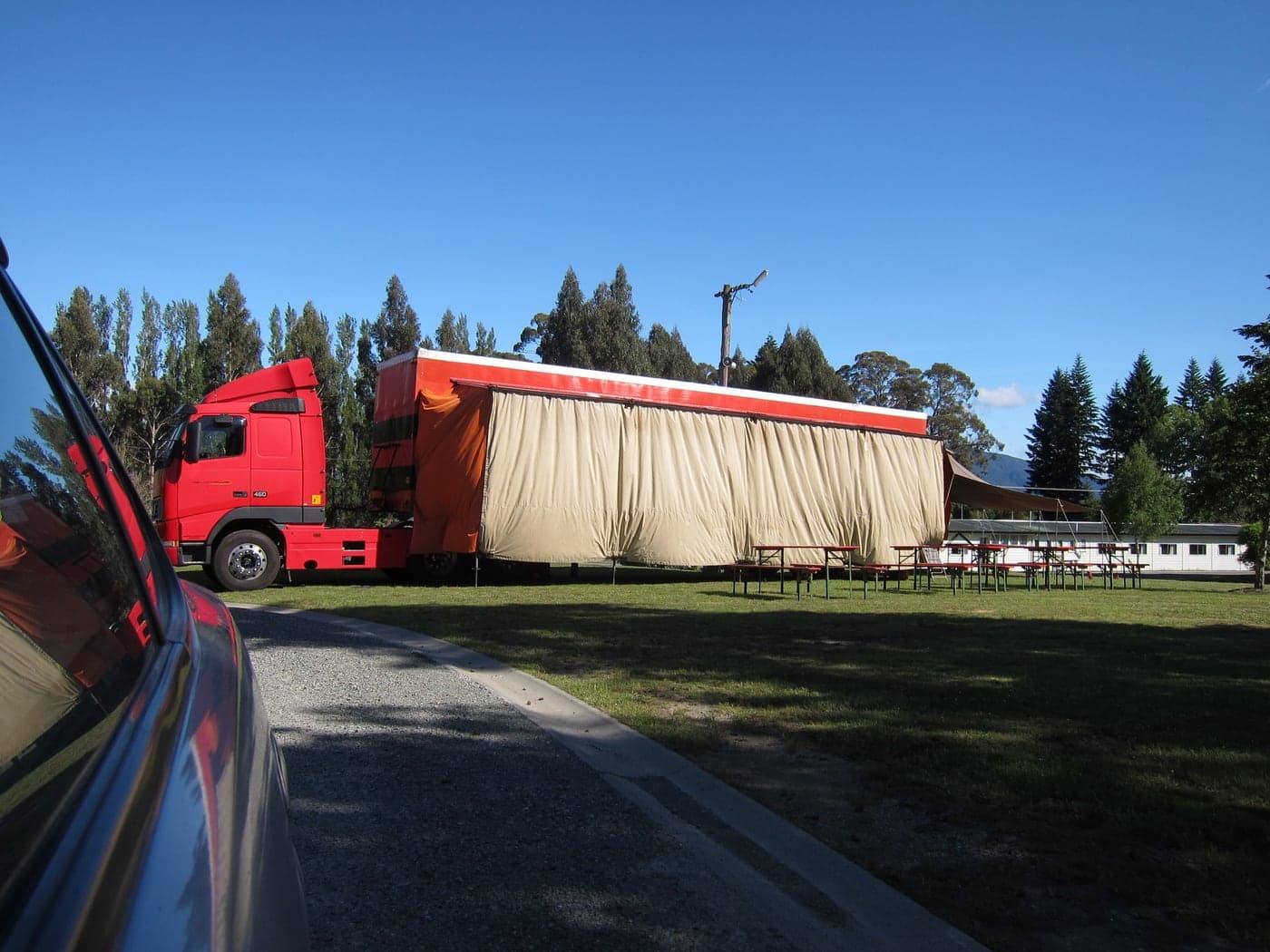 A red touring sleeper vehicle with a large awning beside picnic tables on a grass camping area, with campground buildings and tall trees behind.