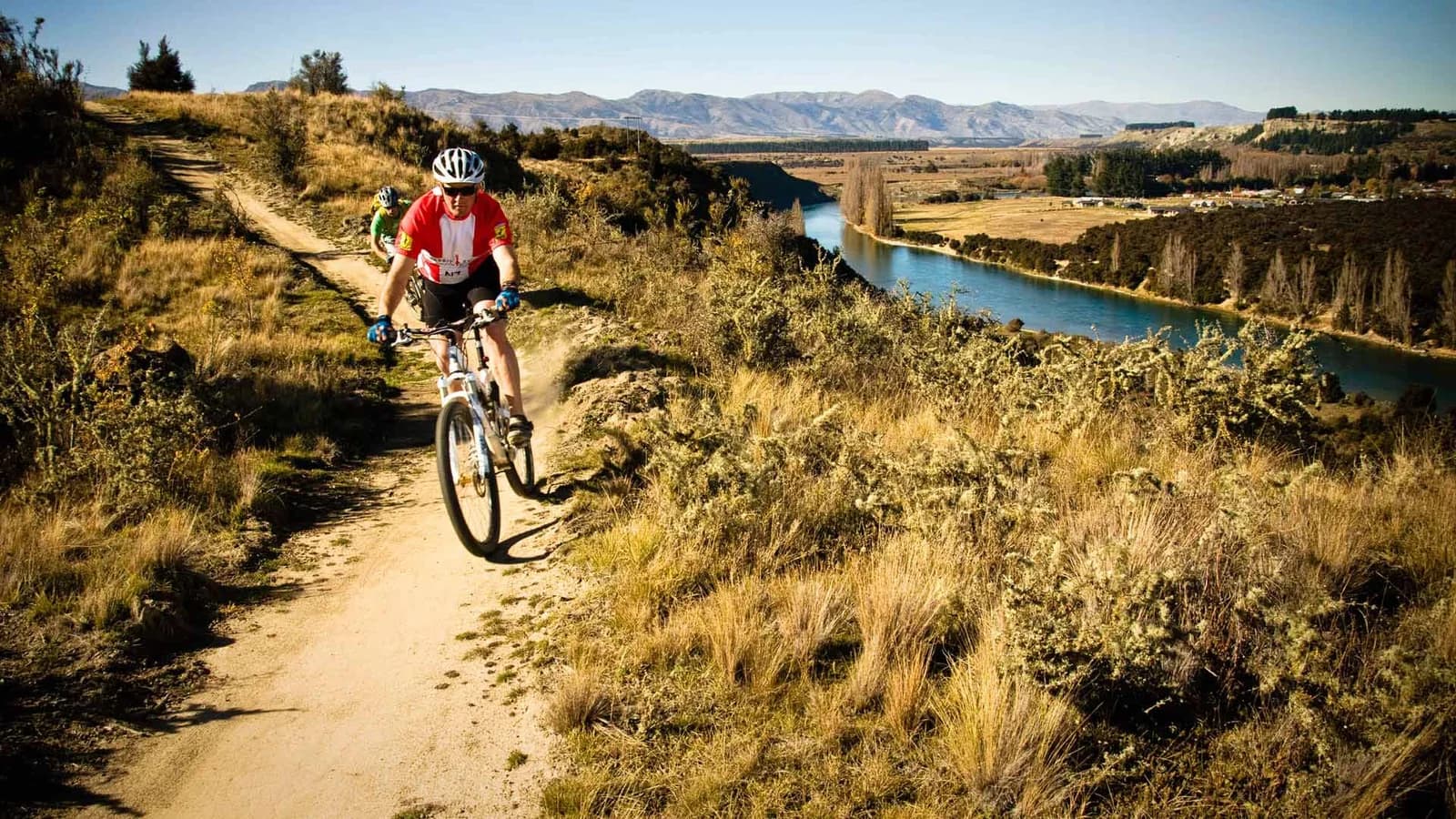Two helmeted cyclists ride a narrow dirt trail above a river, with dry grass and mountains beyond.