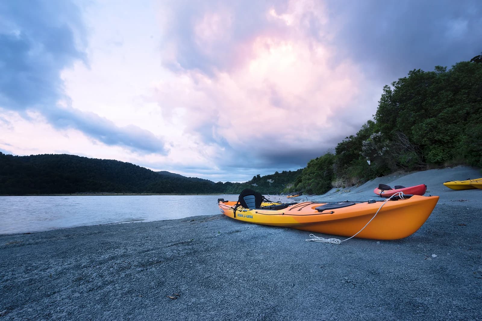 Orange, red and yellow kayaks pulled onto a gravel lakeshore beneath wooded hills and a cloudy sky.