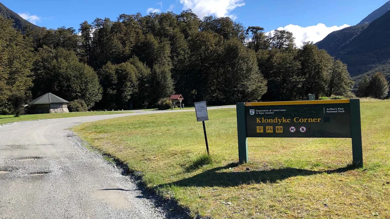 Klondyke Corner sign beside a gravel entrance road, grass and shelter beneath forested mountains.