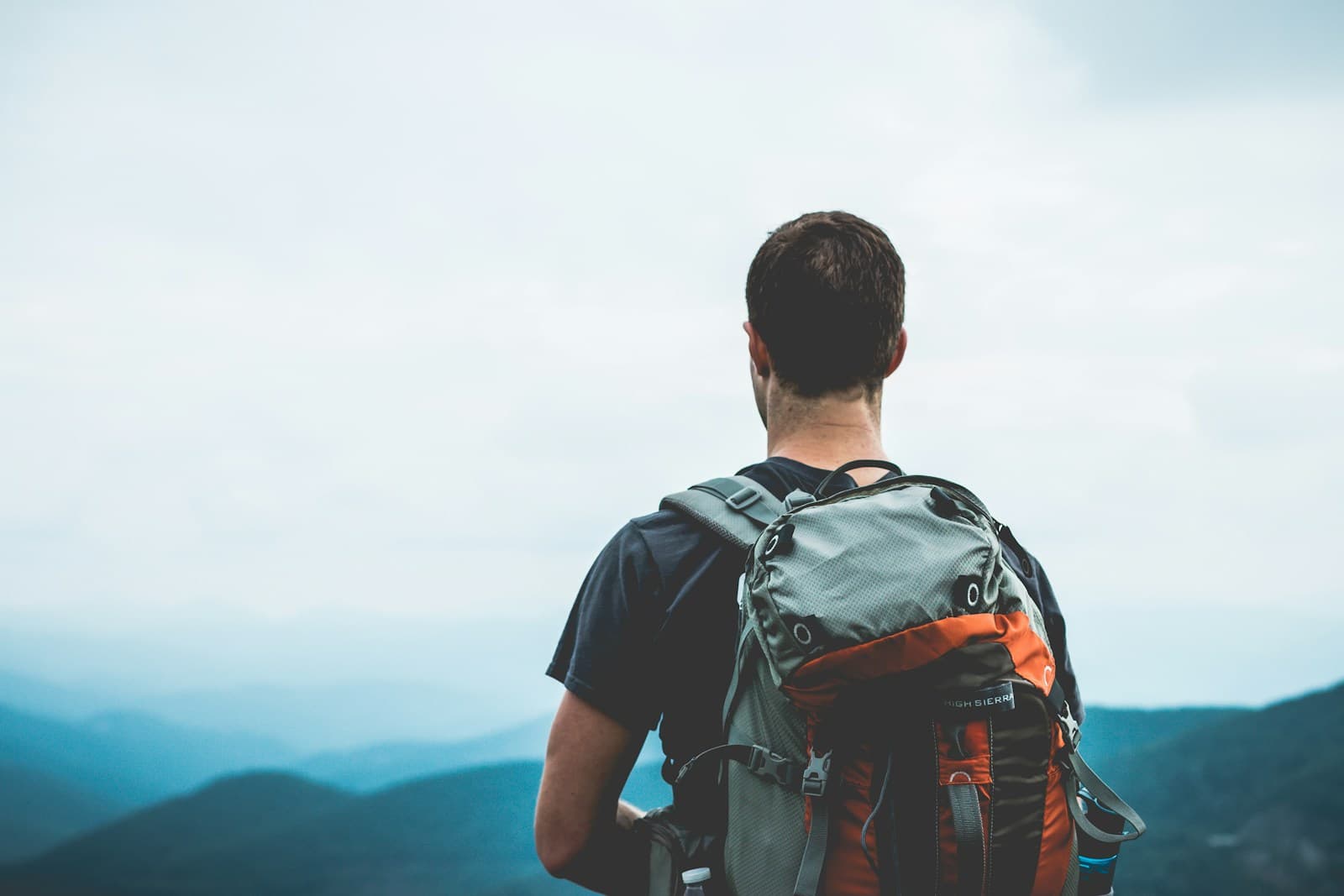 A walker seen from behind carrying a grey and orange backpack, with hills in the background.