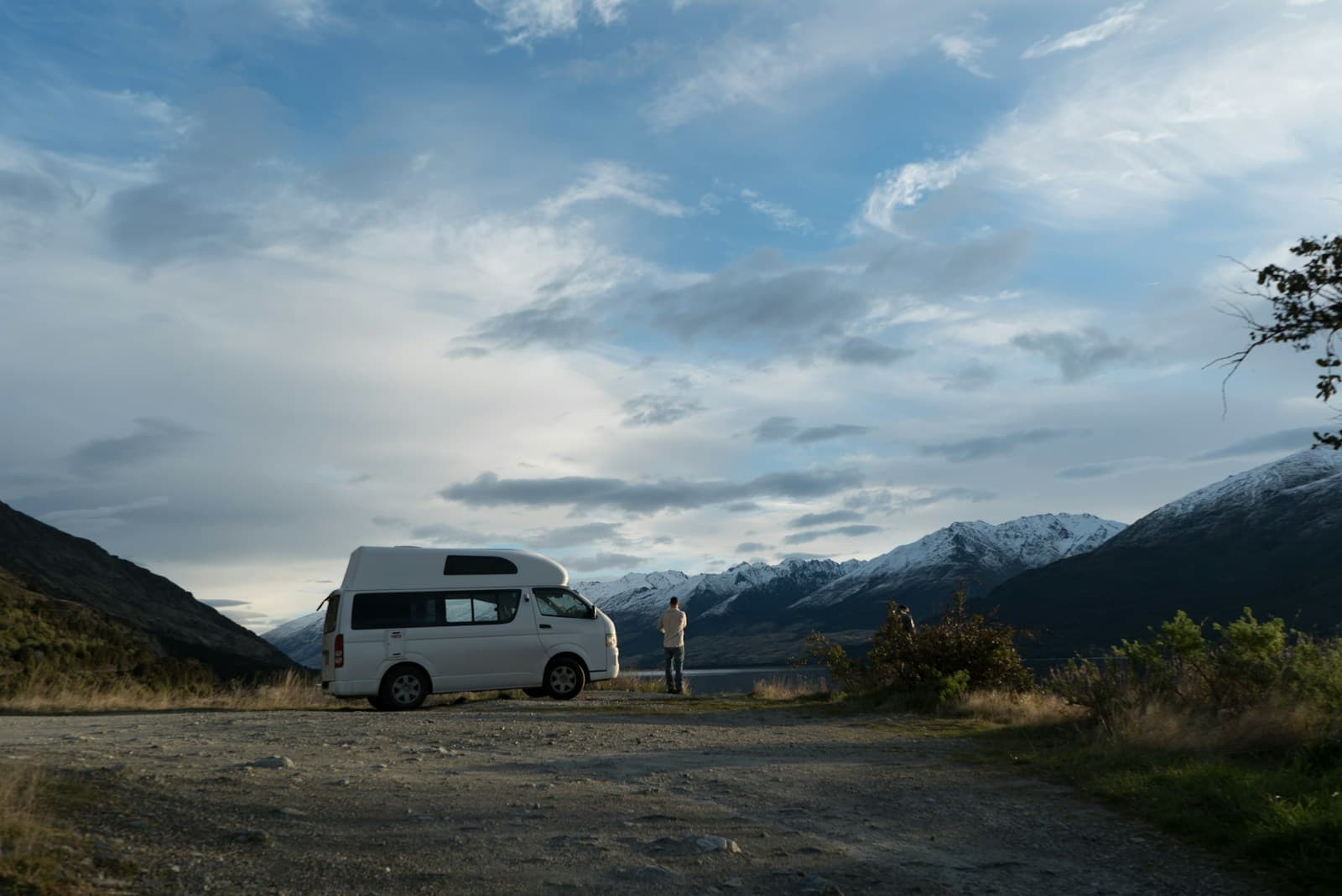 A white campervan and a person beside a lake, with snow-covered mountains in the background.