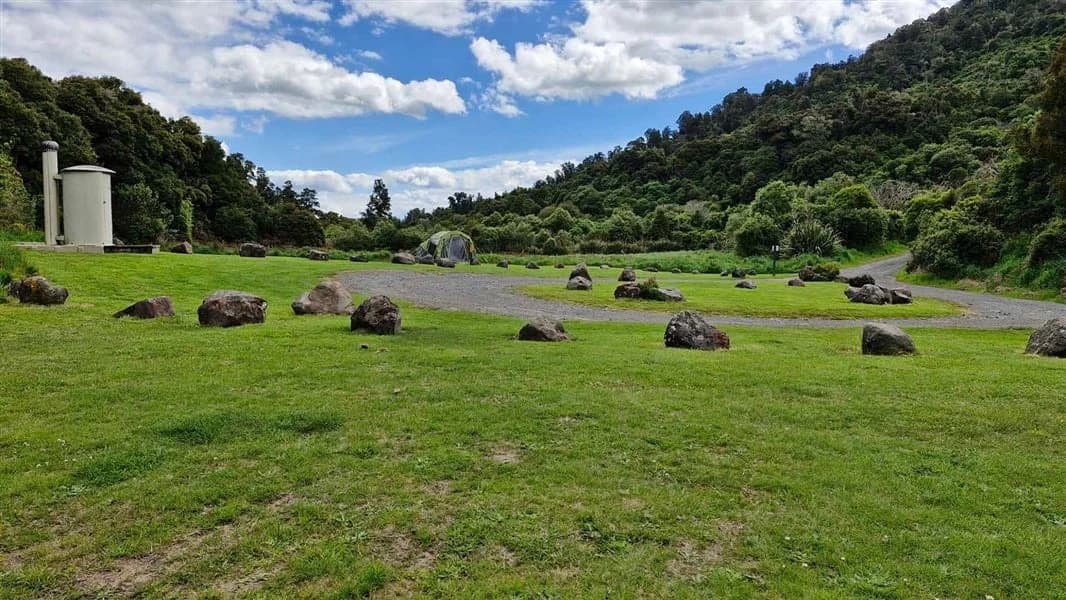 Grass camping spaces separated by large rocks, with a small building and forest at Mangawhero Campsite.