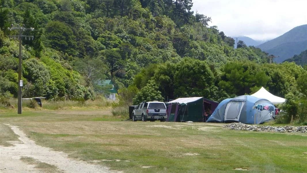 Tents, a camping vehicle and open grassy pitches at Whatamango Bay Campsite beneath forested hills.