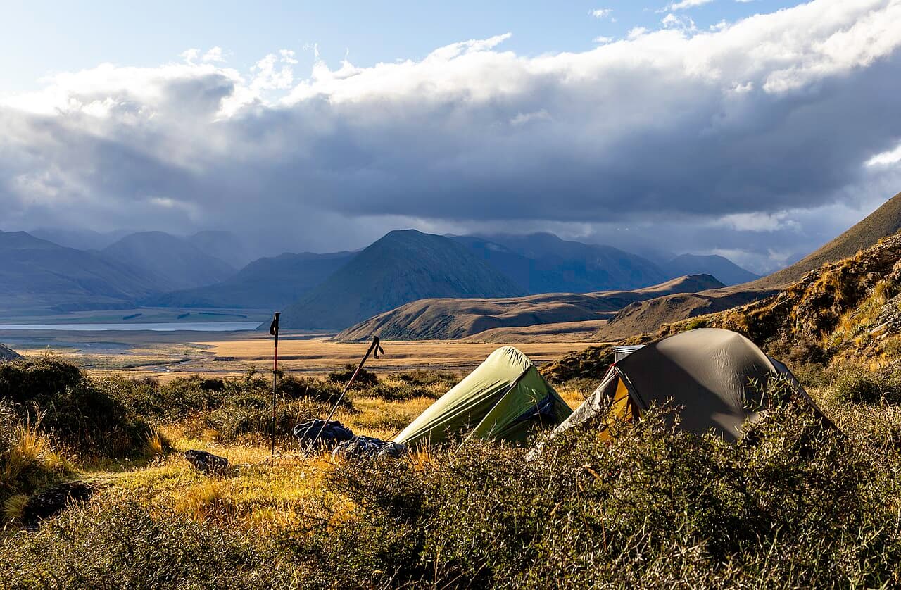 Two tents and trekking poles on grassy ground, with a lake, mountains and clouds in the background.