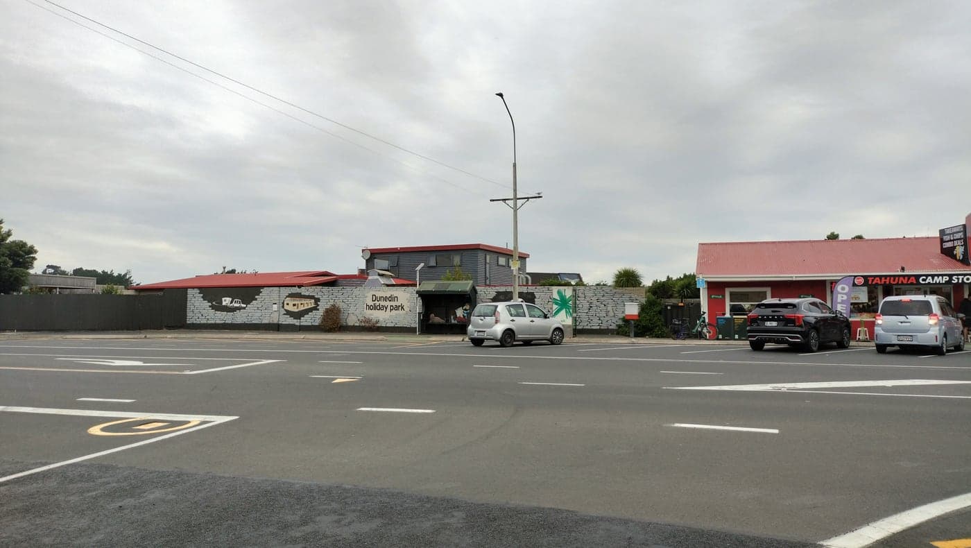 Dunedin Holiday Park's painted frontage and the neighbouring camp store beside Victoria Road, with parked cars.
