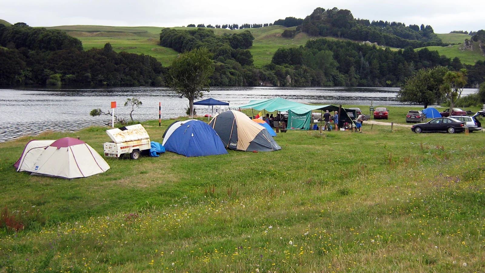 Tents, awnings and parked vehicles on grass beside the water at Arohena Campsite.
